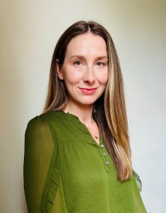 A white woman in a green blouse with shoulder length dark blond hair, freckles, and green eyes smiles softly.